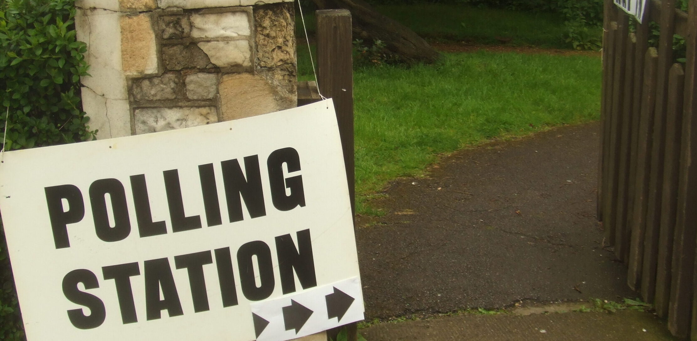 A sign bearing the words 'Polling Station' hangs from a stone gatepost on the edge of a churchyard. The gatepost has a decorative pointy top, signs of wear and exposed brickwork. Arrows on the bottom of the sign point down the path into the churchyard. A wooden gate stands open, inviting the viewer inside.