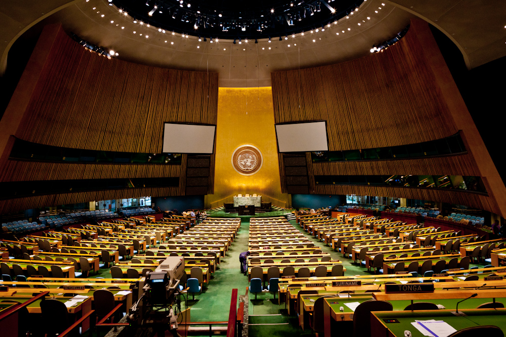 A large room with a high ceiling and no natural lighting. Rows of desks face a podium at the far end of the room. The names of some UN member states are visible on some desks in the foreground. Much of the interior decor is golden and the UN logo is prominently placed in the centre of a large gold-coloured coloured pillar behind the podium.