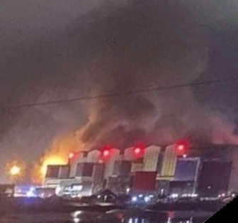 A large industrial building at night, seen at a distance over water, with a very large smoke cloud rising above it.