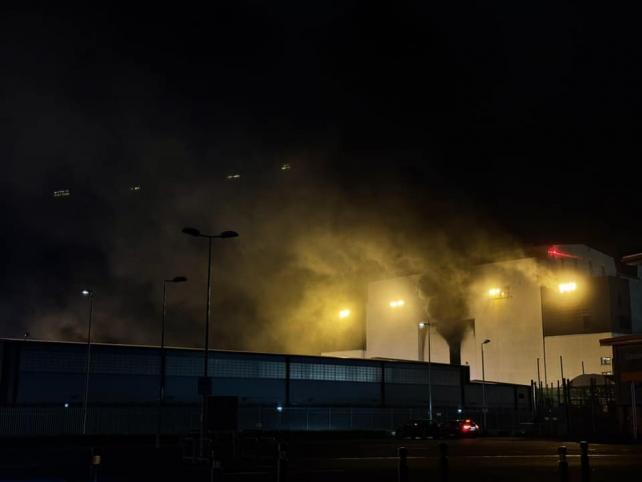 Black smoke rises from the front and roof of a very large industrial building at night time. Smaller buildings are silhouetted in the foreground. The only illumination is the orange lights from the building.