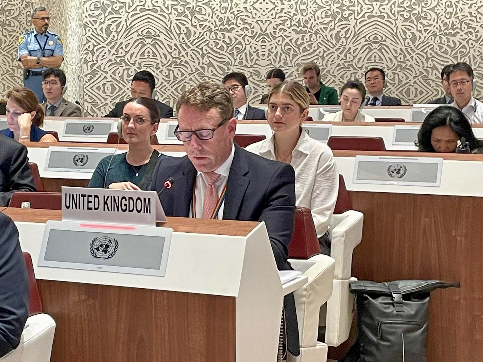 A man in a suit, sitting down at bench bearing the UN logo and the name 'United Kingdom' is speaking into a microphone. Behind him are several rows of benches with the UN logo and smartly dressed people sitting behind them. Most people are wearing distinctive ear pieces for simultaneous translation.