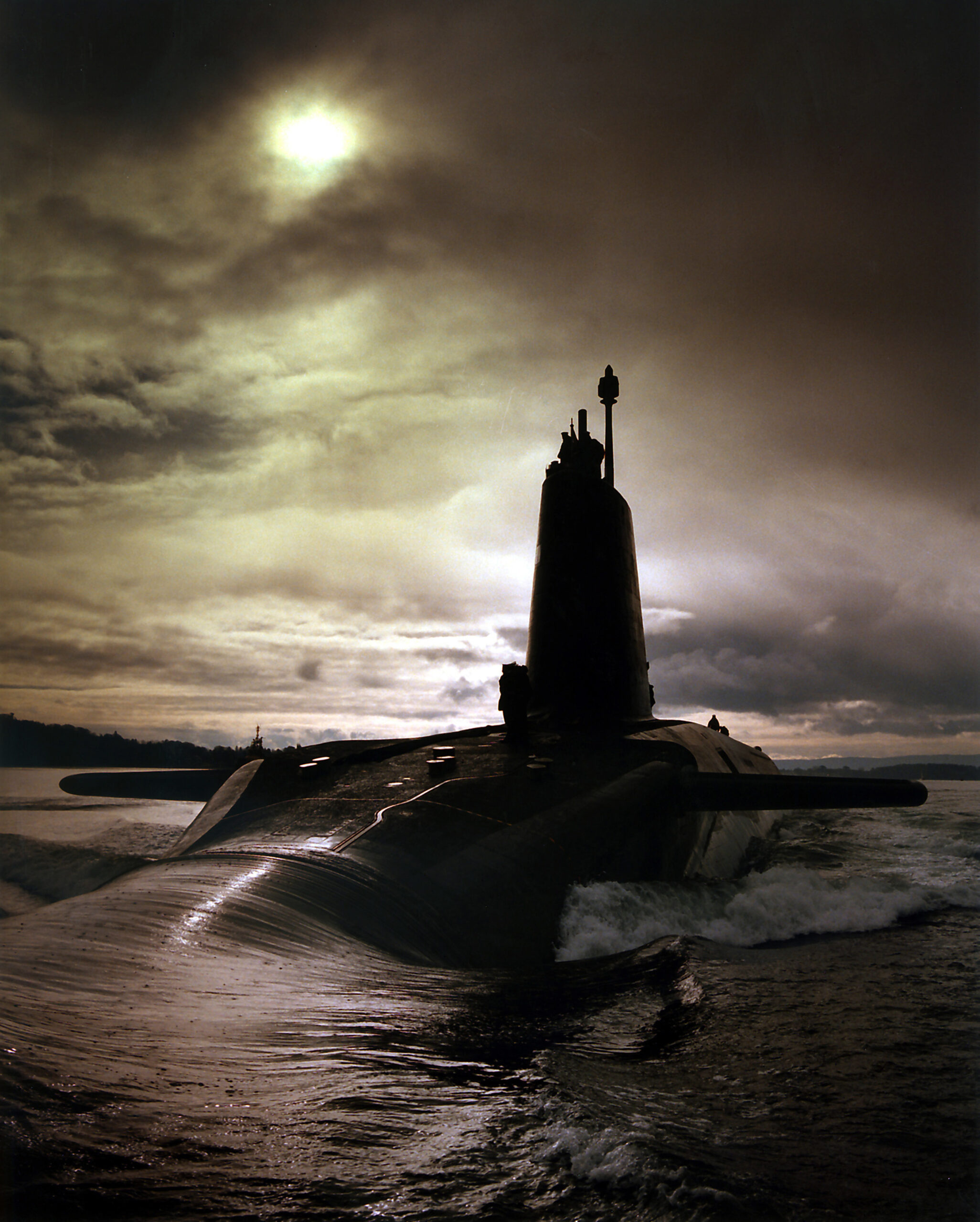 Water rushes over the front of a silhouetted submarine, in front of a cloudy sky.