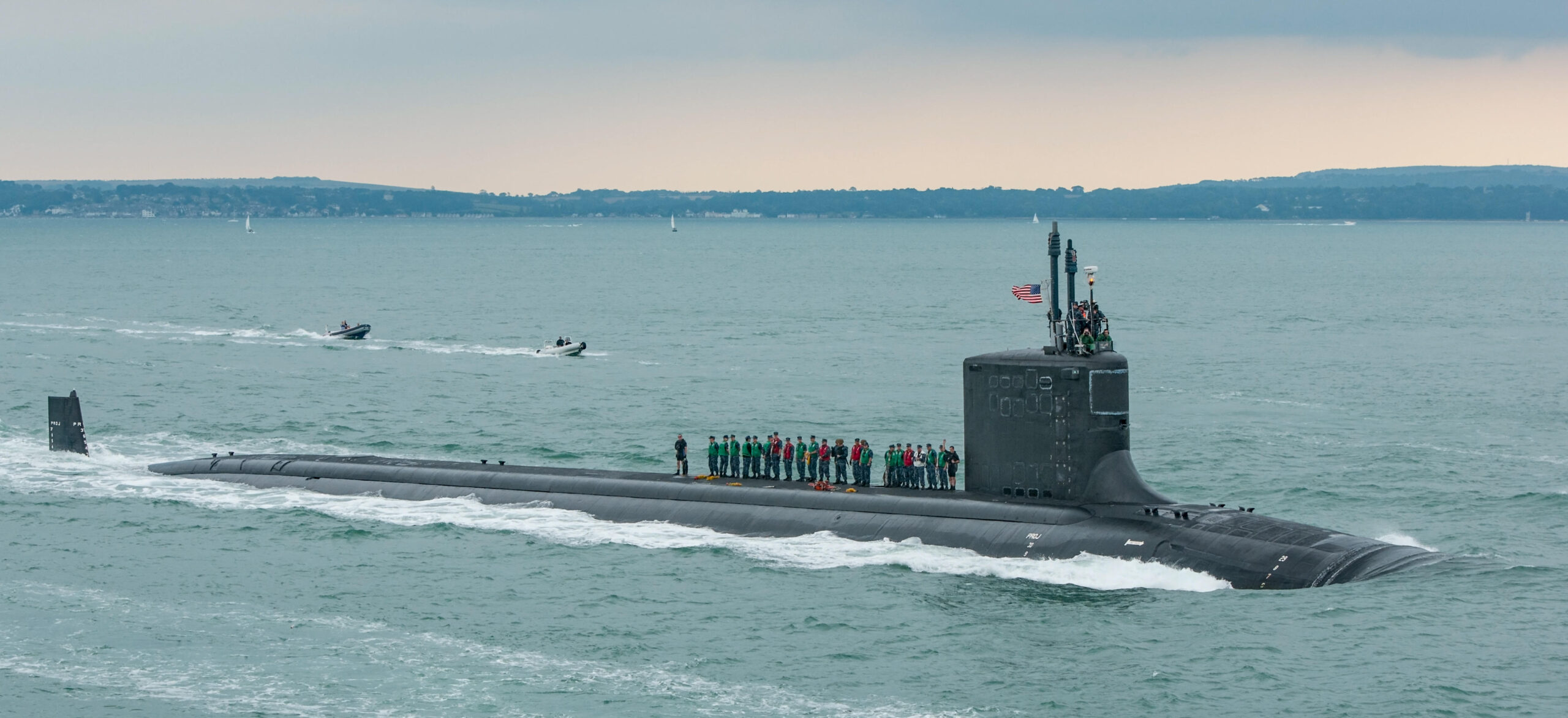 A grey submarine moves through the water with a US flag flying from its fin. Shoreline is visible in the background, and many of the crew are standing on the submarine hull, wearing life jackets. Two rigid inflatable boats follow close behind the submarine.